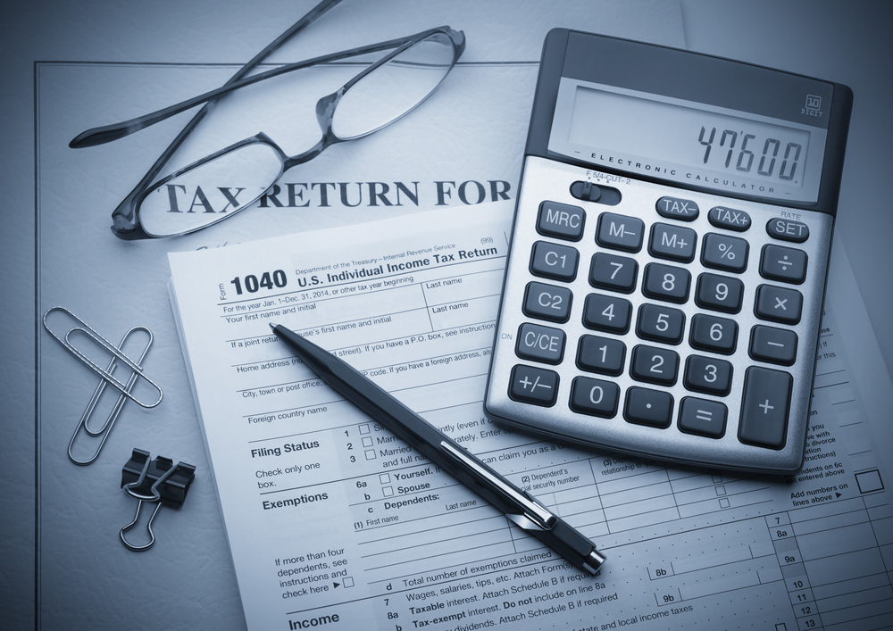 A calculator sitting on top of tax forms on a wooden table with a hand holding a pen nearby.