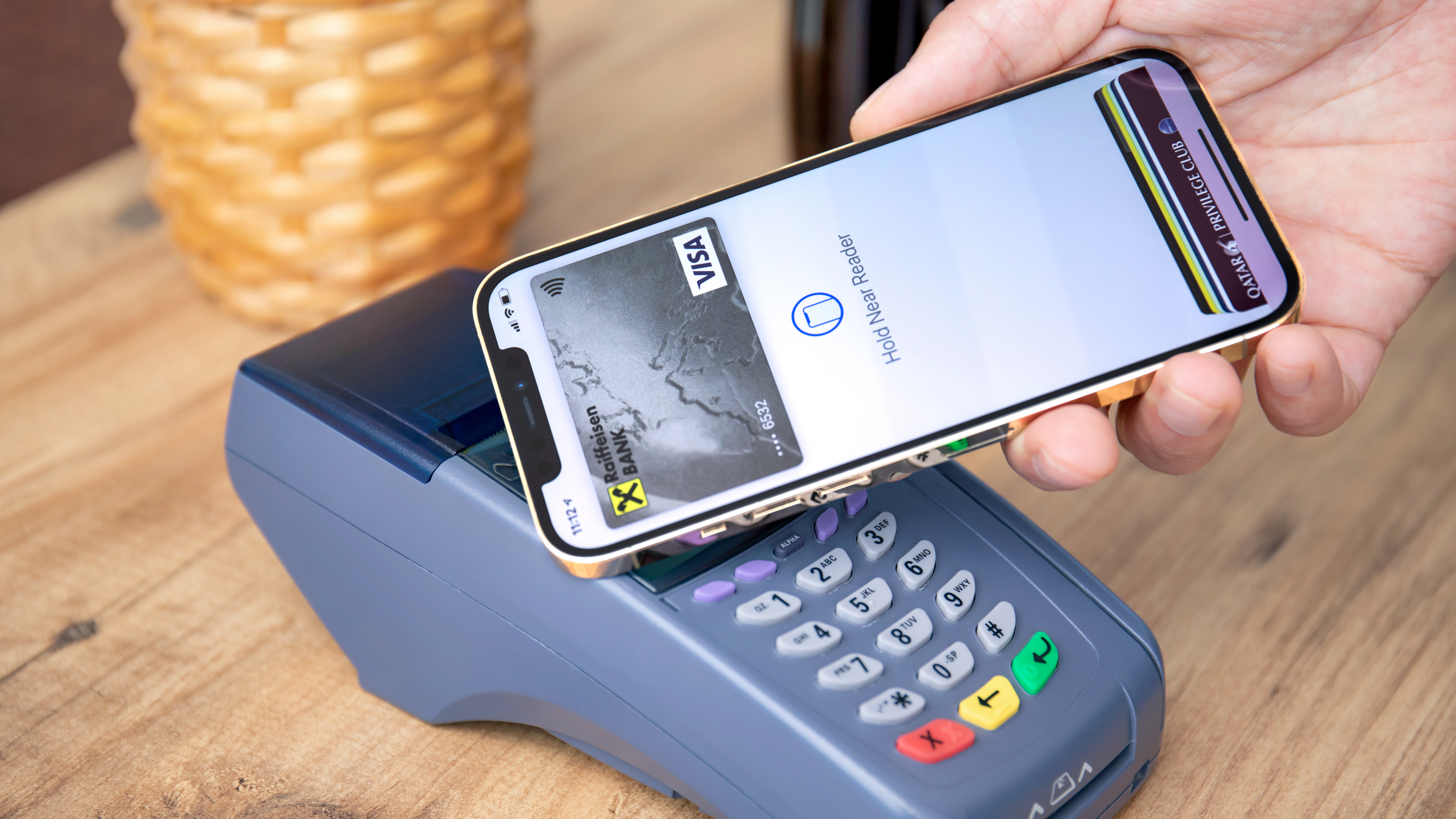 A close-up of an iPhone screen showing the Wallet app with a bank card selected, held in a shopper’s hand near a store checkout counter