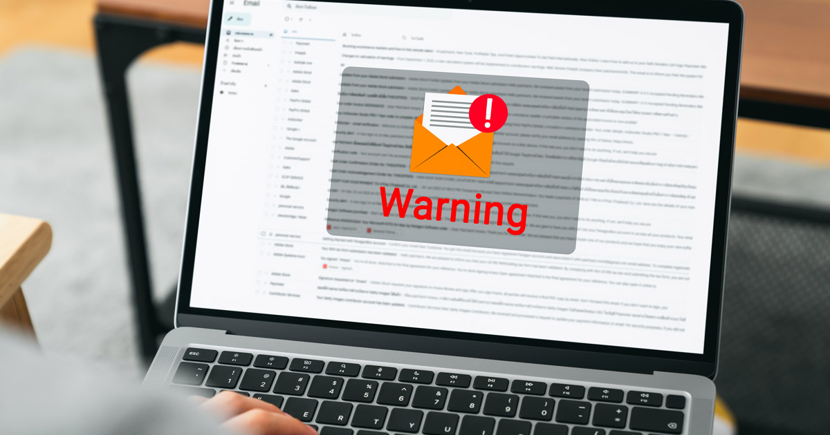 A close-up photo of an open laptop showing an email inbox with a suspicious message highlighted, hands hovering over the trackpad as if deciding whether to click