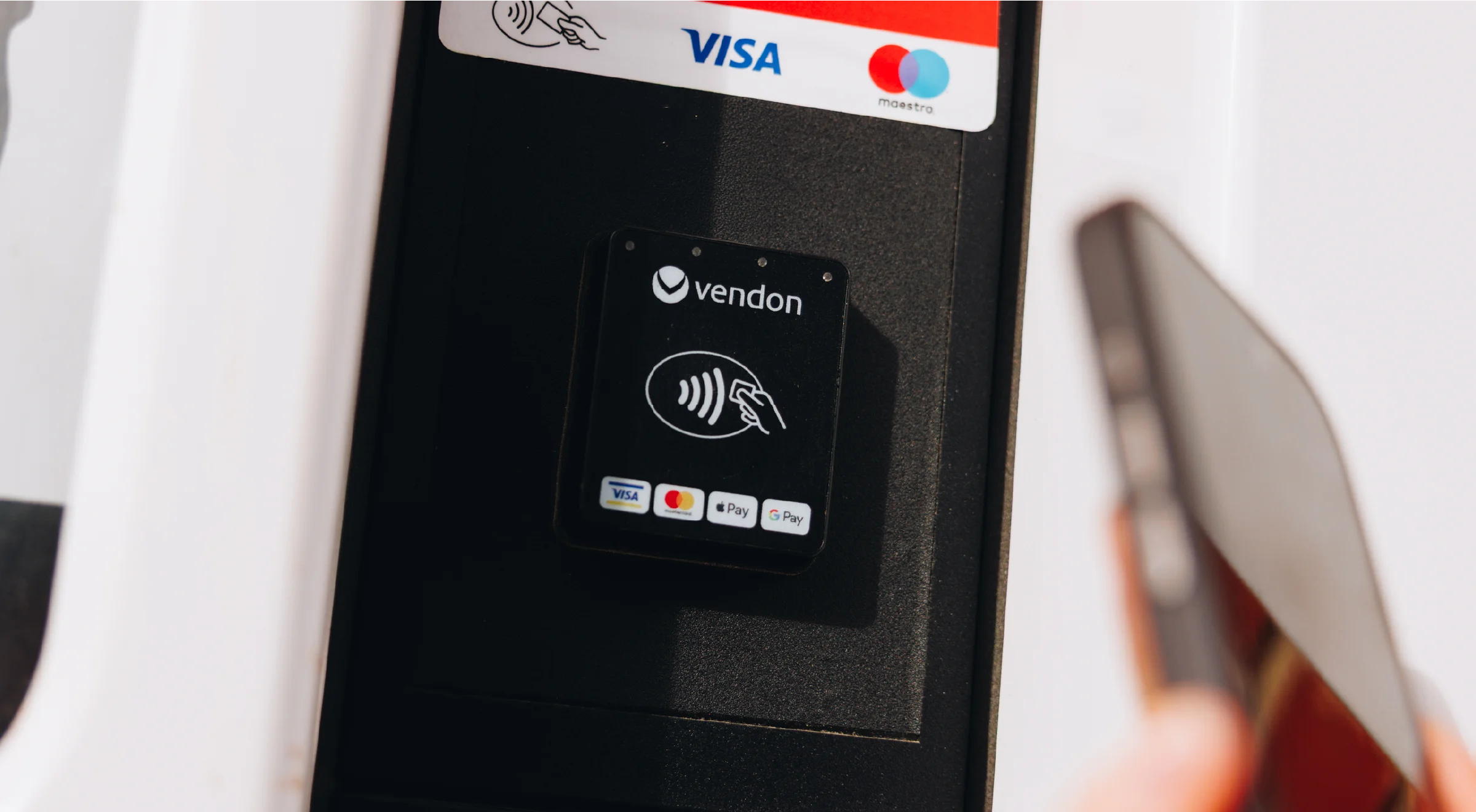A contactless-enabled payment terminal on a retail counter with a shopper’s hand holding a credit card and a smartphone nearby