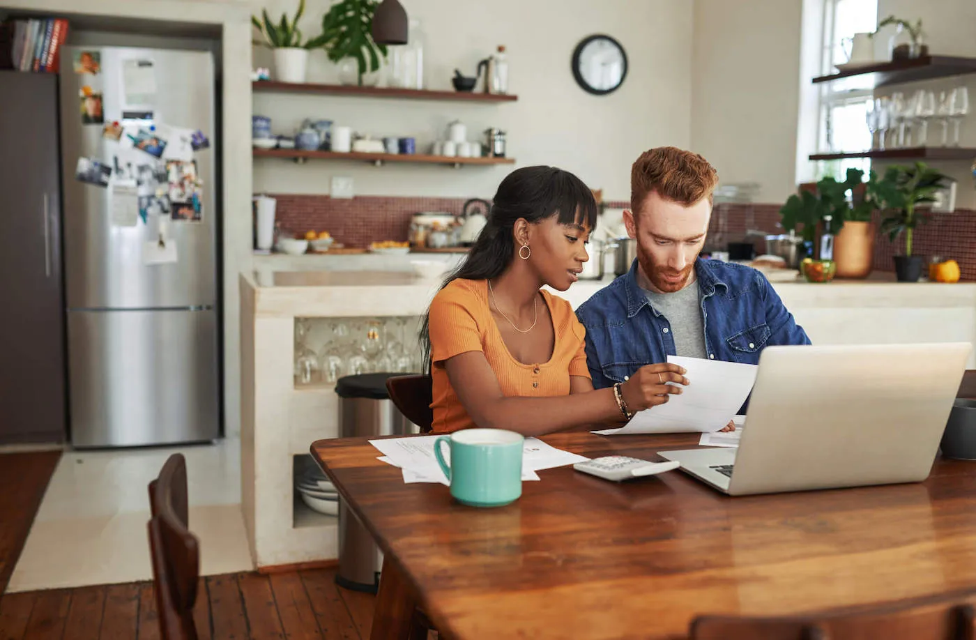 A couple sitting at a kitchen table reviewing mortgage paperwork with a calculator and laptop, natural indoor light, real-life photo