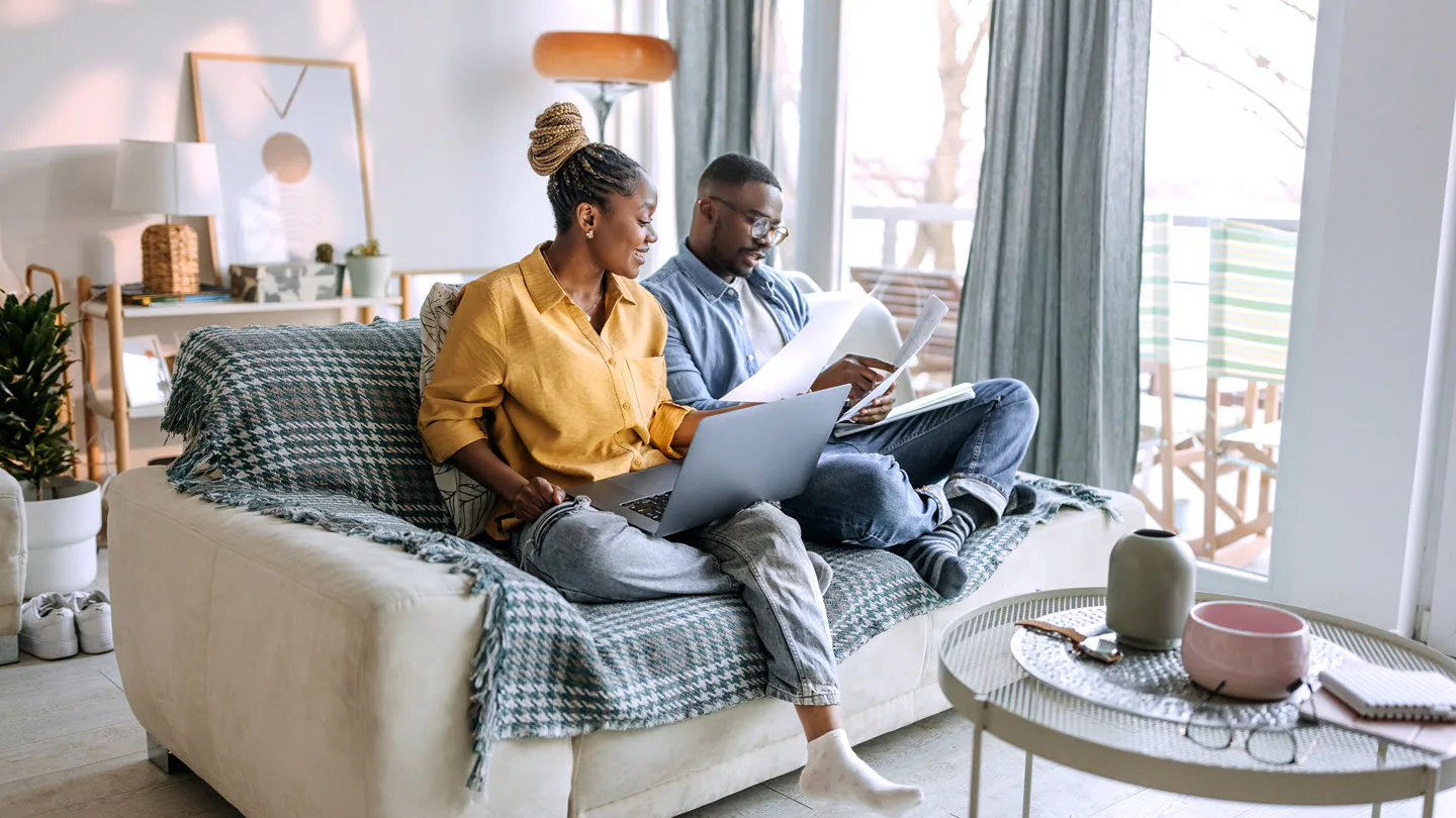 A couple sitting on a couch at home with a laptop open and a notebook, smiling while reviewing a simple budget together, natural candid photo