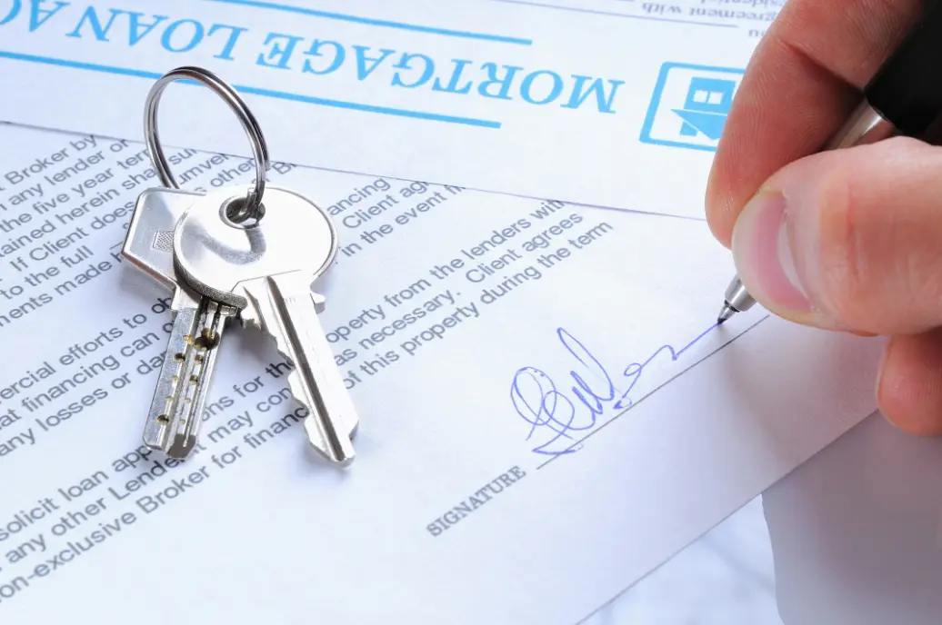 A first-time homebuyer sitting at a kitchen table signing mortgage documents with a loan officer, natural window light, real photography style