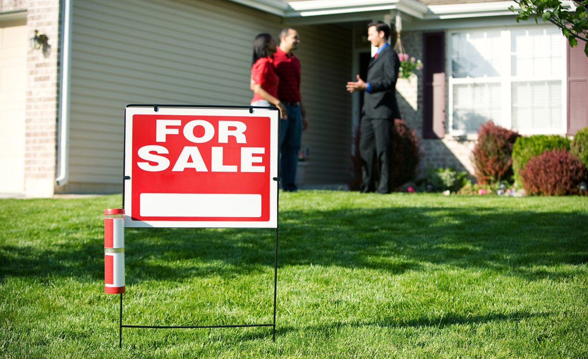 A for-sale sign in the front yard of a home, with paperwork and a pen visible on a nearby porch table