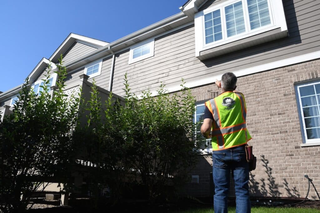 A home appraiser standing in a driveway taking photos of a house exterior with a tablet, daytime real estate photography style