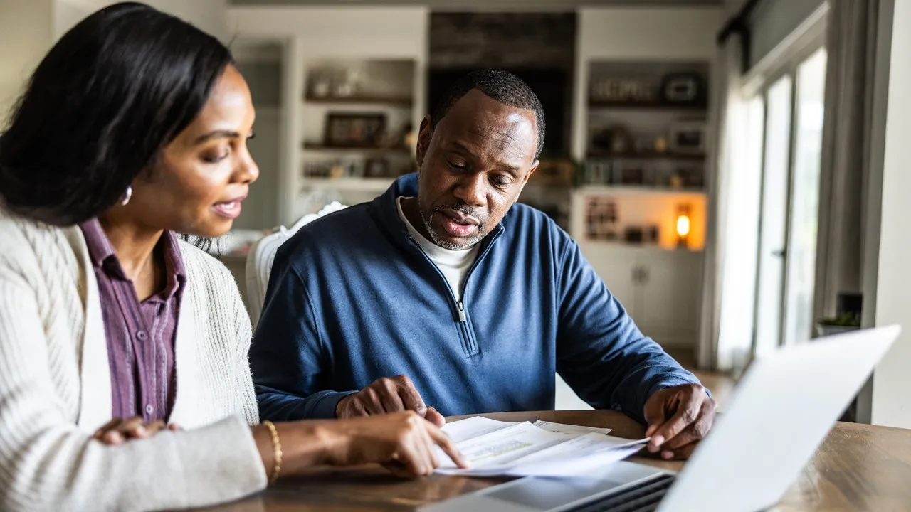 A homebuyer sitting on a couch using a laptop and reviewing printed loan estimates on a coffee table, warm indoor lighting