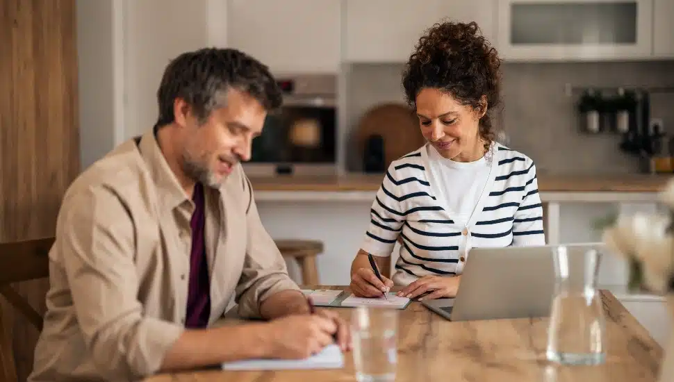 A married couple sitting at a kitchen table at night reviewing paper tax forms and a laptop, with a calculator nearby, real photography style