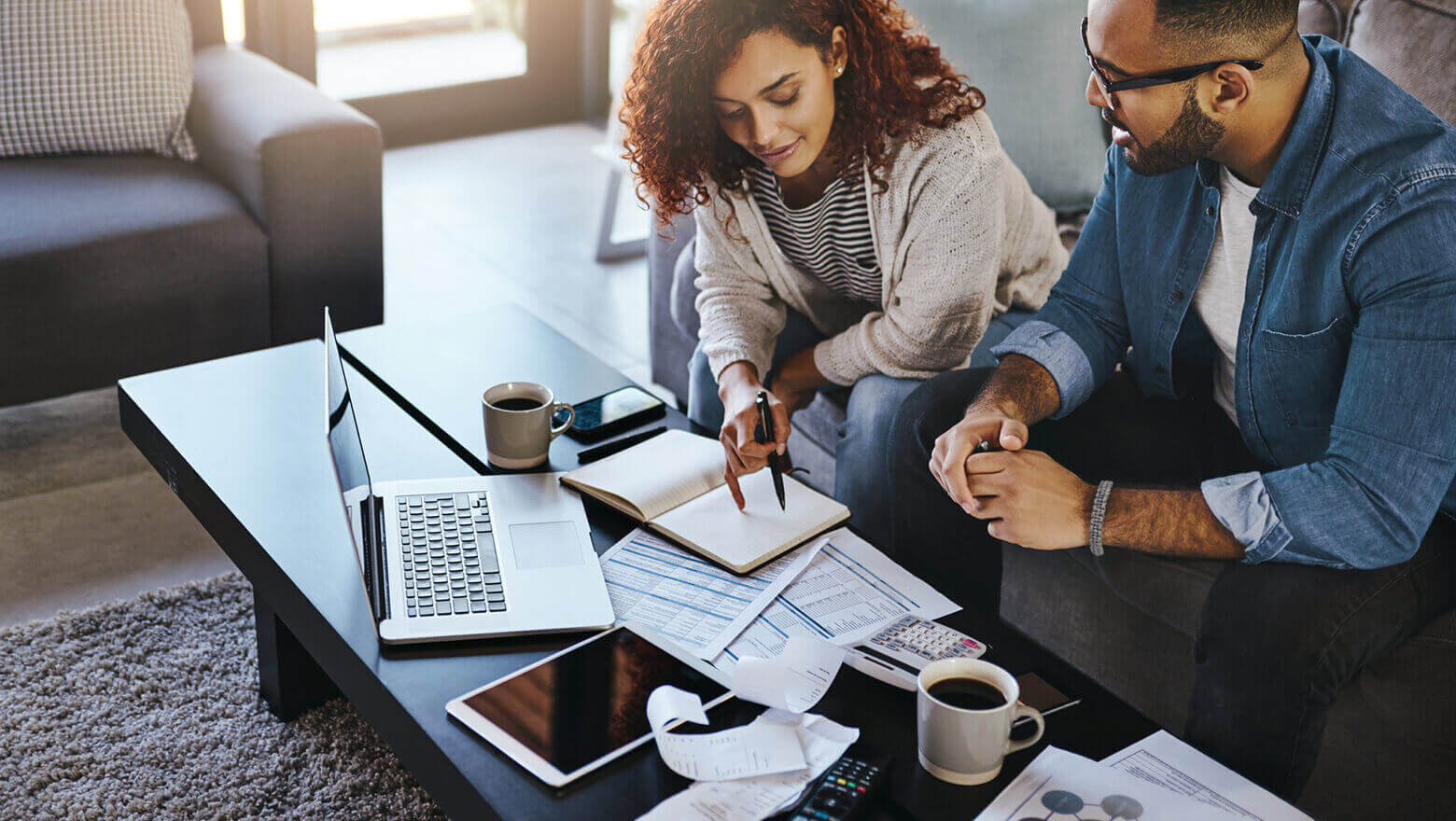 A married couple sitting on a living room couch reviewing pay stubs together with a laptop open on a coffee table, candid photo