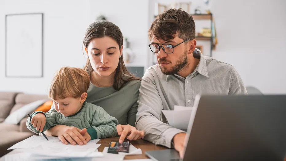 A mother and father sitting on a couch with a laptop open, talking through a household budget and savings goals, candid lifestyle photo