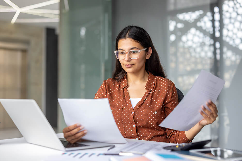 A person at a desk using a laptop and a calculator while reviewing printed health insurance plan details, realistic home office photo