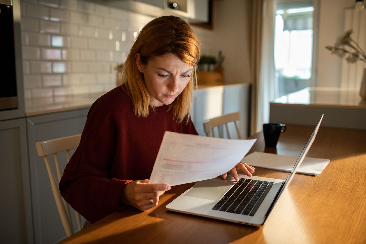 A person at home using a laptop to pay household bills with paper statements nearby on a tidy desk, realistic lifestyle photo