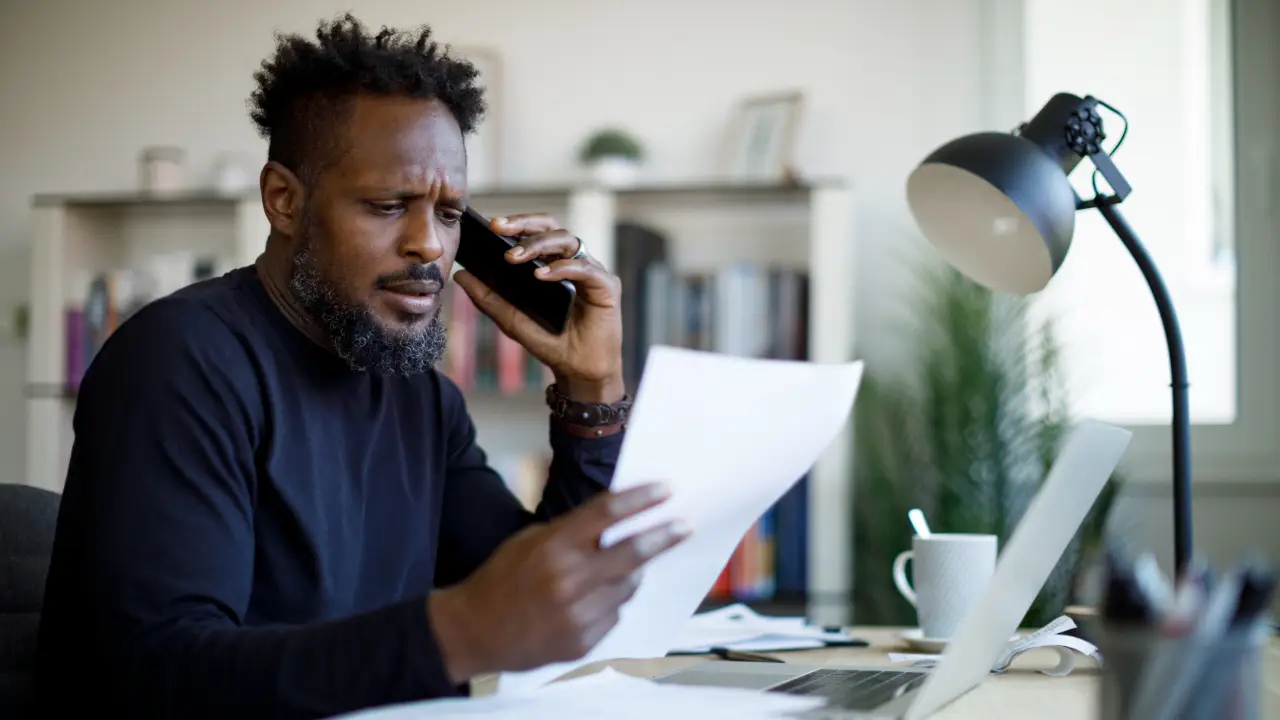 A person in a home office holding a phone while reviewing printed closing documents on a desk, realistic photo