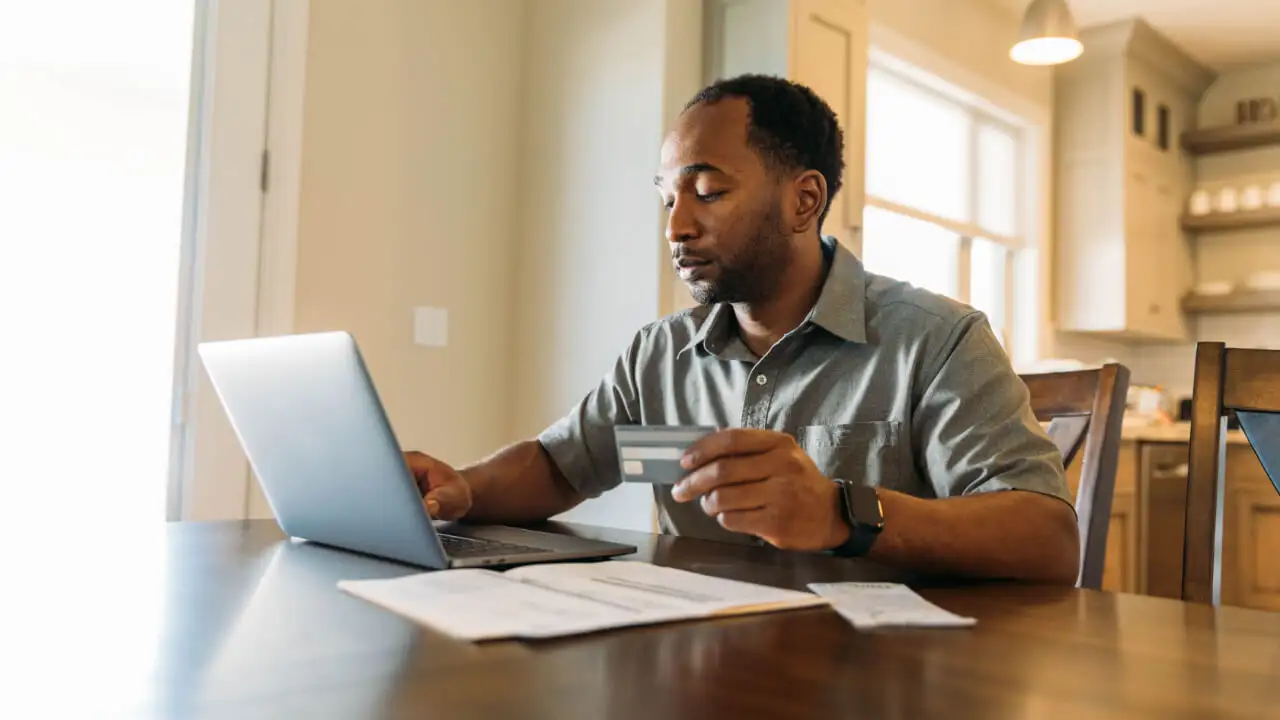 A person making an online credit card payment on a laptop at a dining table with a credit card and notebook nearby, real-life photo