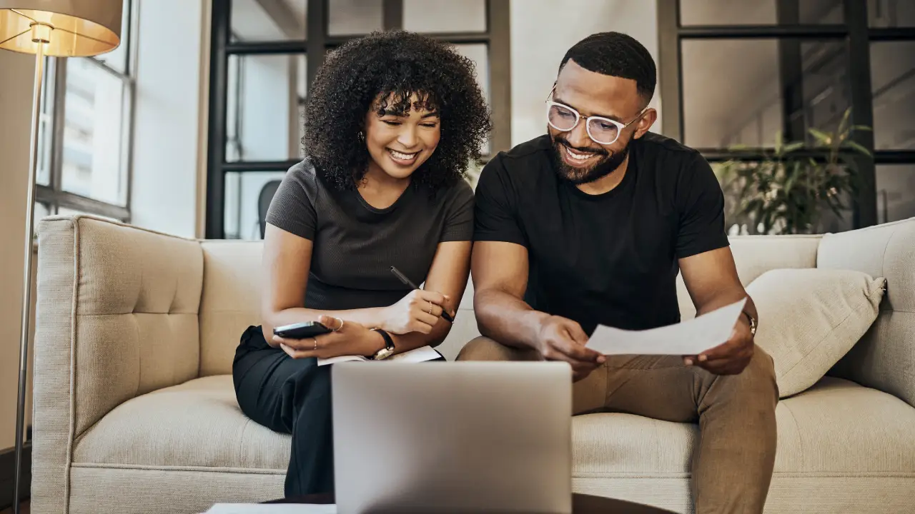 A person relaxing on a couch holding a smartphone open to a banking app, with a notebook on the coffee table, everyday personal finance moment
