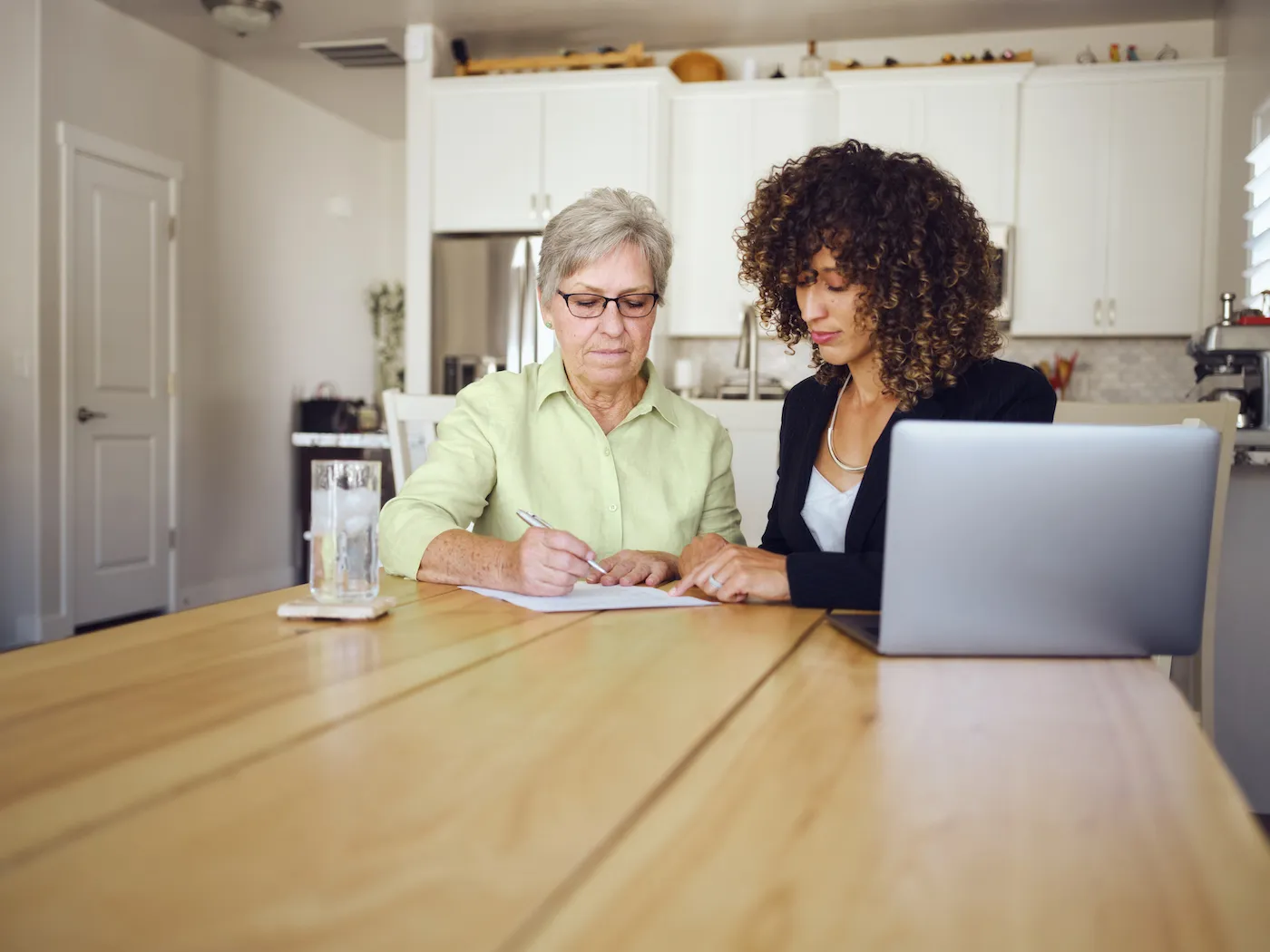 A person signing loan paperwork at a kitchen table with another adult nearby reviewing documents, real-life personal finance moment