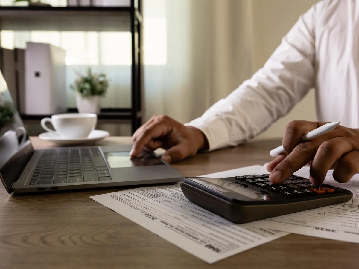 A person sitting at a home office desk using a calculator beside tax forms and a laptop, realistic documentary photography