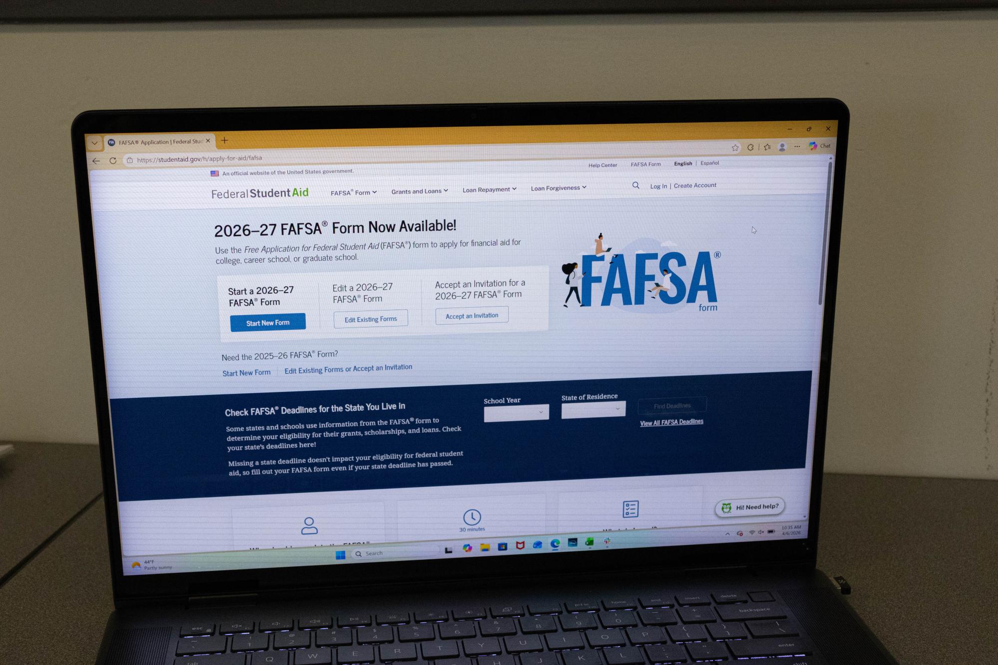 A person sitting at a kitchen table using a laptop to log into the Federal Student Aid website, with paperwork and a notebook nearby, candid photo