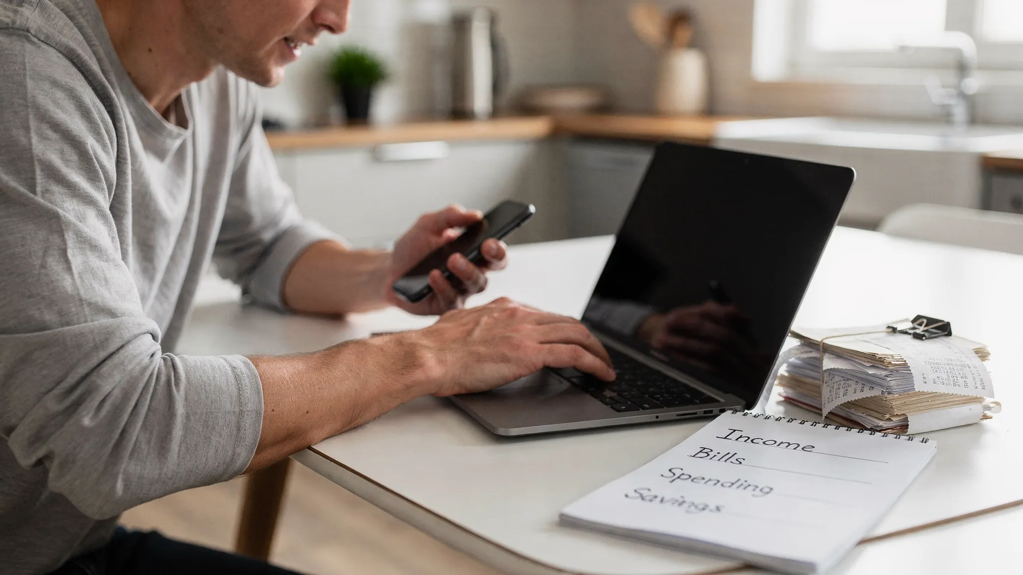 A person sitting at a kitchen table with a laptop open and a notebook beside a cup of coffee, reviewing a monthly budget in a calm home setting, real photograph style