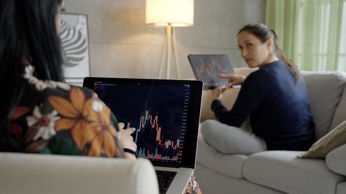 A person sitting on a couch at home holding a smartphone open to a brokerage investing app, with a laptop on a coffee table, warm indoor lighting, candid photo