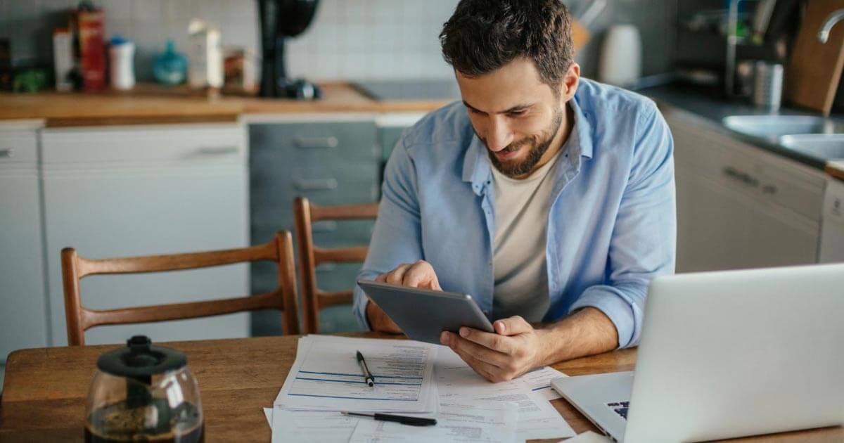 A person using a calculator and reviewing car payment paperwork at a kitchen table with a laptop open, natural daylight photography style