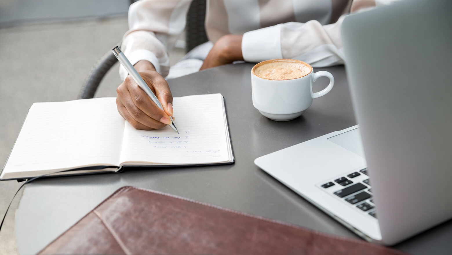A person writing a simple budget in a notebook next to a bank card and a cup of coffee on a wooden table, realistic photo