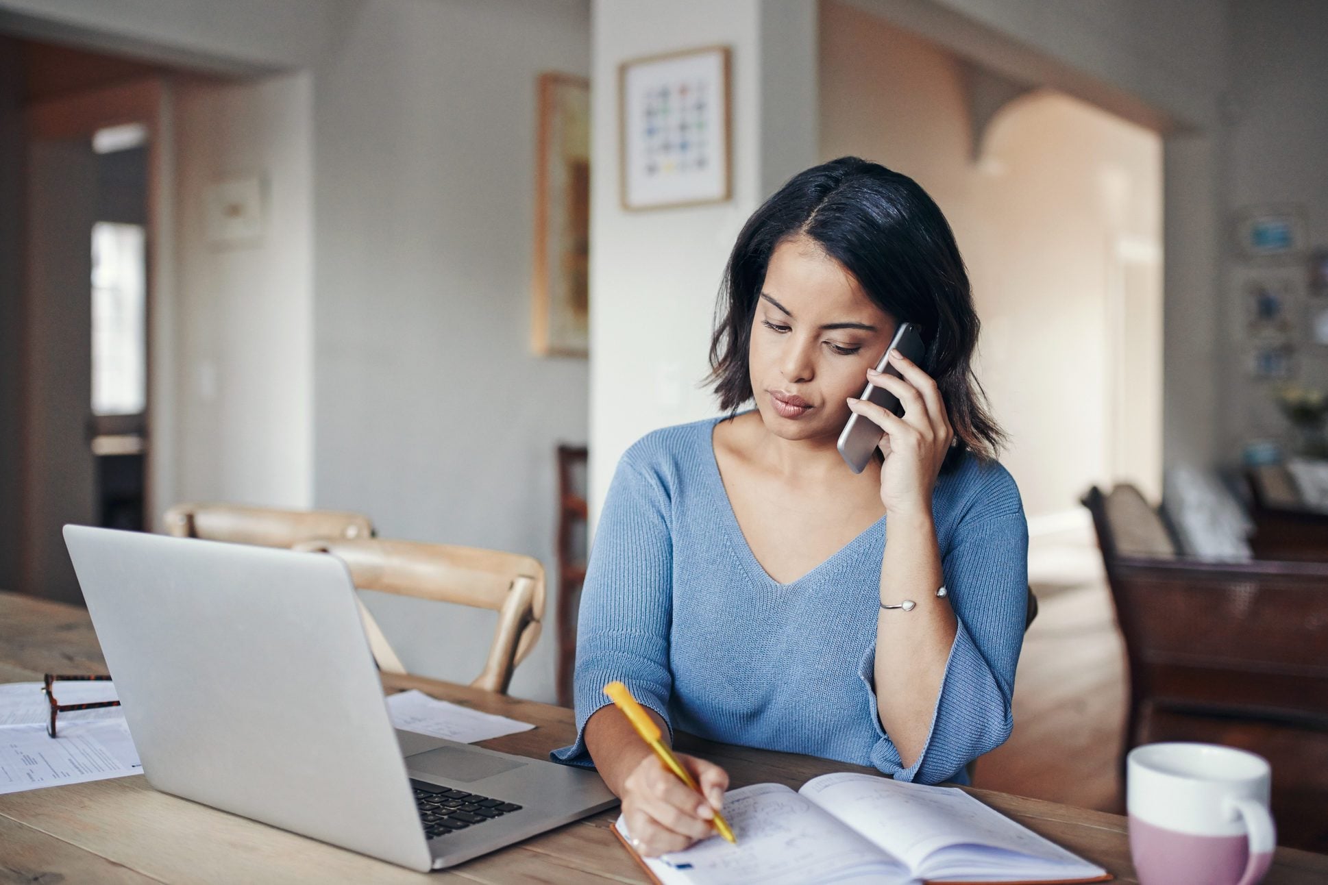 A person writing notes in a notebook next to a printed bill and a laptop on a dining table, realistic photography