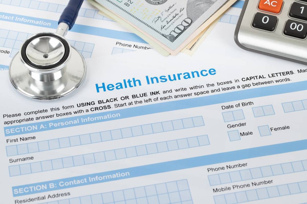 A person’s hands using a calculator next to health insurance paperwork and a notebook on a dining table