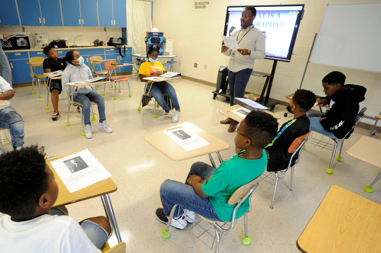 A public school teacher sitting at a classroom desk after hours, grading papers with a laptop open and a coffee mug nearby, realistic indoor photography