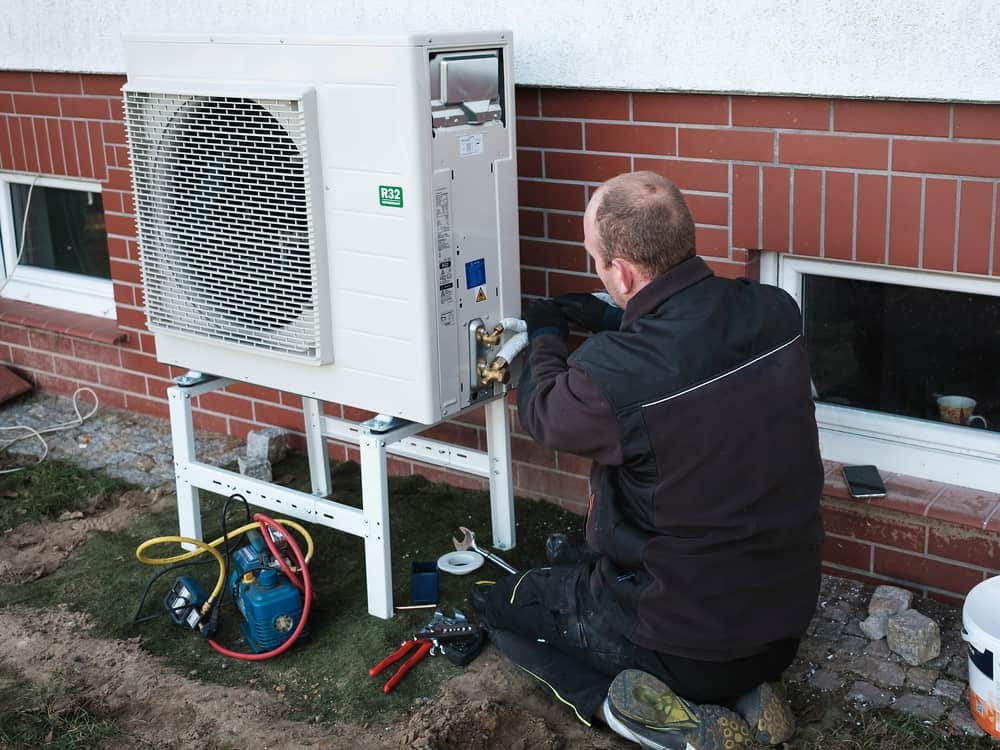 A residential HVAC technician installing an outdoor air source heat pump unit next to a single-family home on a clear day, real photo