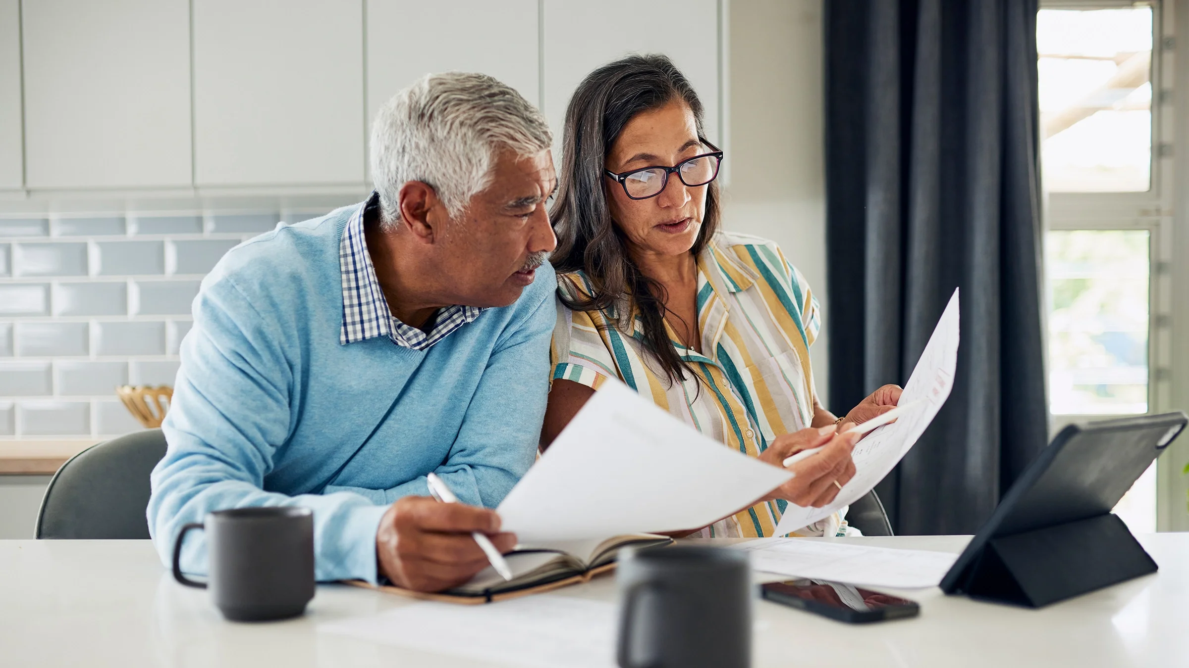 A retired couple sitting at a dining table reviewing health insurance documents and a laptop together, candid home photo