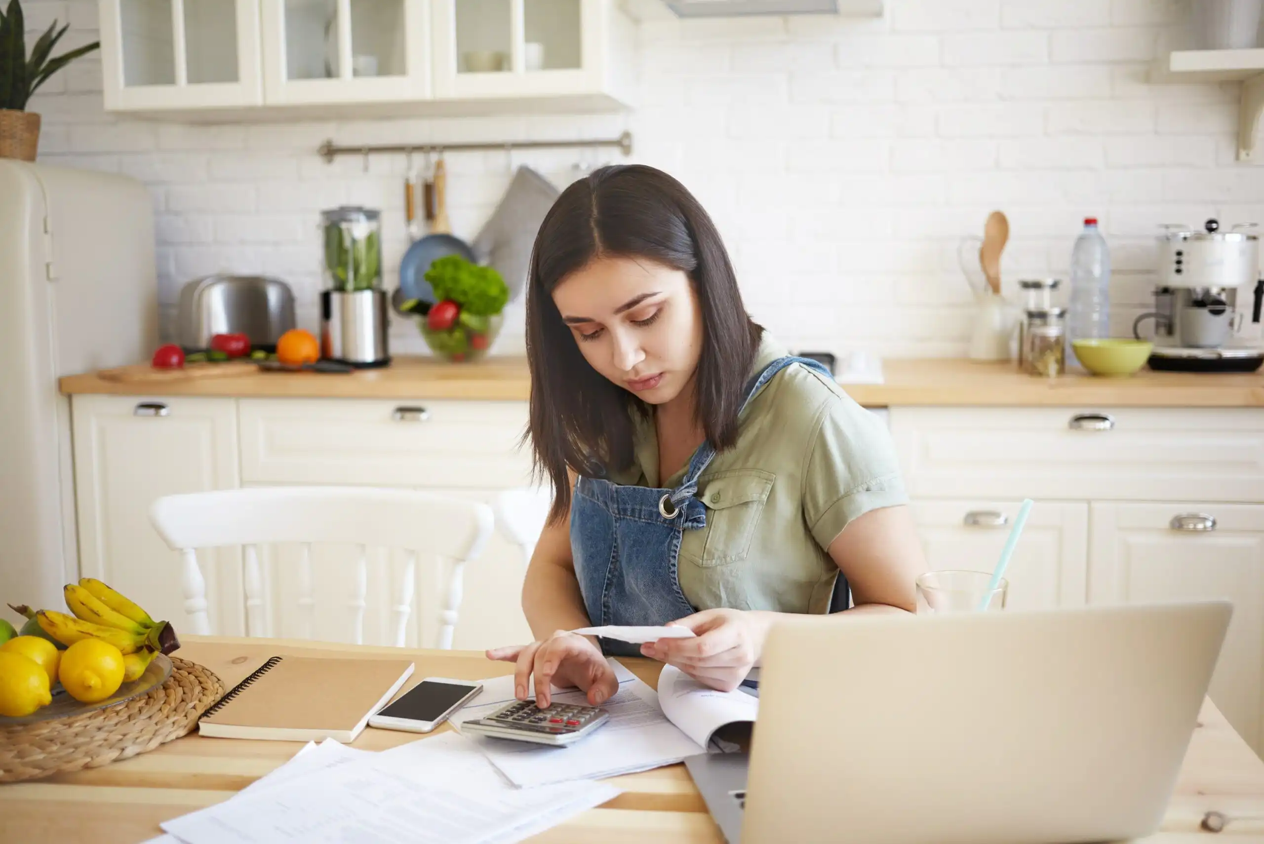A woman sitting at a kitchen counter using a laptop with a budgeting spreadsheet open, realistic candid photo