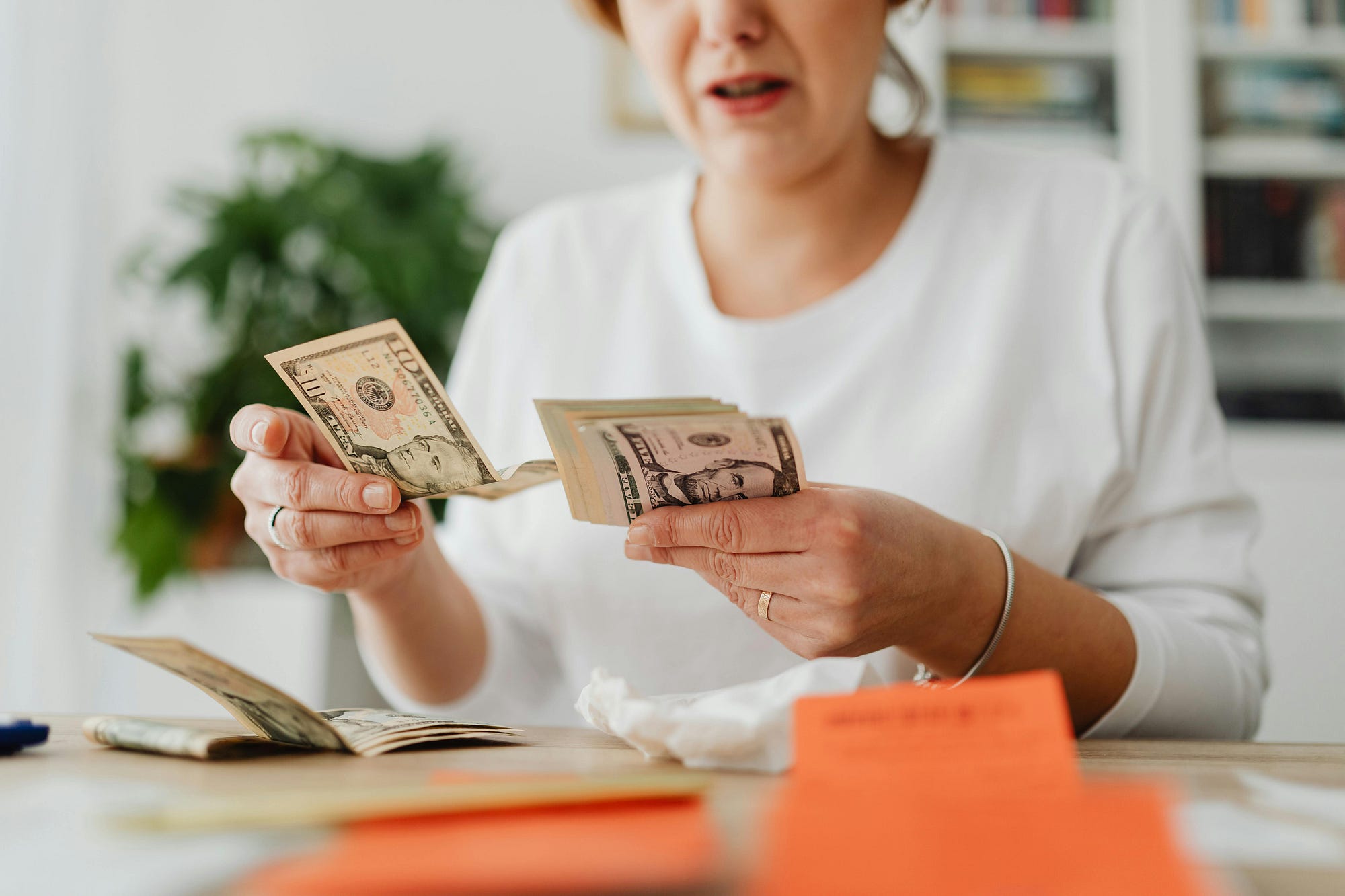 A woman sitting at a kitchen table counting cash next to a stack of bills and a smartphone, realistic candid photo