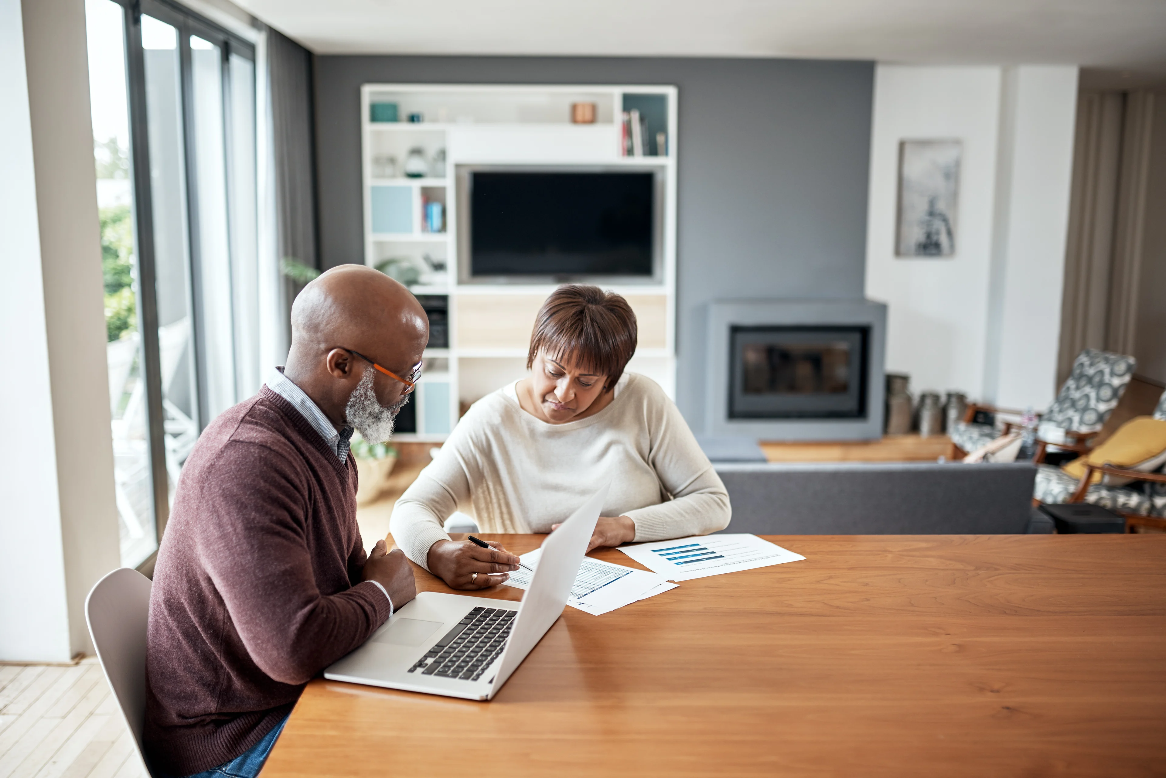 An older couple sitting at a kitchen table reviewing bills and a laptop budget spreadsheet in a warm home setting